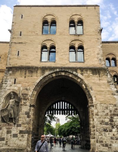Ein großes historisches Stadttor aus Stein mit drei Torbögen und Bogenfenstern, auf dessen linker Seite die Statue einer Person in Rüstung steht, markiert den Beginn vieler Kölner Stadtführungen. Menschen gehen und radeln hindurch, im Hintergrund sind Bäume und Gebäude zu sehen.